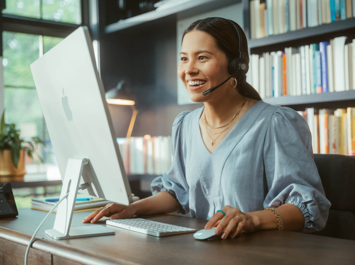 A woman smiling, wearing a headset and working at a computer.